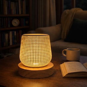 May include: A glowing, cylindrical lamp with a dotted pattern sits on a wooden base. The lamp emits a warm, inviting light. The lamp is on a dark wooden table, next to a book and a mug. The background includes a bookshelf and a couch.