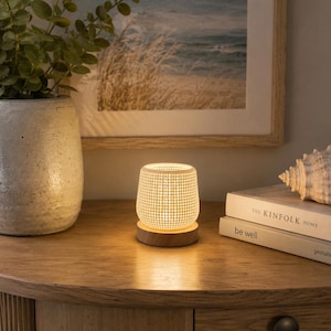 May include: A small, cylindrical white lamp with a perforated grid pattern sits on a circular wooden base. The lamp emits a warm, soft glow, illuminating the surface around it. The design features a repeating dot texture across the exterior.