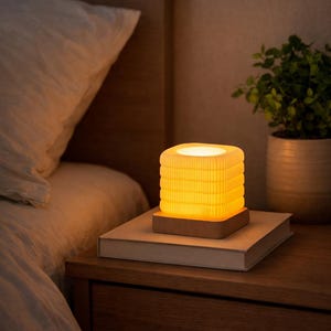May include: A glowing, square-shaped bedside lamp with a warm yellow light. The lamp sits on a wooden base, atop a closed book on a wooden nightstand. A white pillow and a potted plant are in the background.