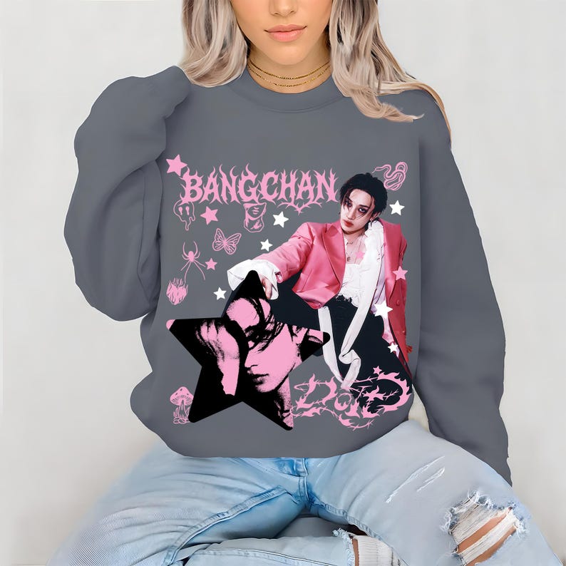 Pode incluir: Camisola cinzenta com um gr&aacute;fico rosa de uma pessoa com um casaco rosa e o texto "BANG CHAN". O design inclui estrelas, uma aranha, uma borboleta e uma imagem em forma de estrela de um rosto. Estilo casual.
