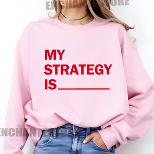 May include: Pink sweatshirt with the text "MY STRATEGY IS" in red.