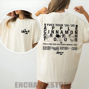 May include: Cream-colored oversized t-shirt featuring a black graphic on the front and back. The back graphic reads "YVES TOUR '24-'25 APPLE CINNAMON CRUNCH TOUR" with cartoon illustrations and tour dates. The front has a smaller logo.