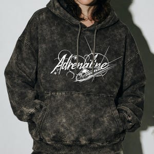 Puede incluir: Una sudadera con capucha gris oscuro con lavado &aacute;cido, con capucha con cord&oacute;n y bolsillo delantero. El gr&aacute;fico blanco en la parte delantera dice "Adrenaline" en una fuente estilizada, con un dise&ntilde;o de tren. La sudadera est&aacute; hecha de un material suave y c&oacute;modo.