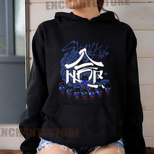 Może przedstawiać: Czarna bluza z kapturem z białym nadrukiem koreańskiego boysbandu Stray Kids. Nadruk zawiera nazwę zespołu po angielsku i koreańsku, a także stylizowany wizerunek członk&oacute;w zespołu.