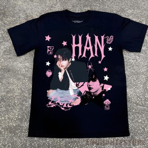 Pu&ograve; includere: T-shirt nera con un design grafico rosa. Il design presenta il nome "HAN" in un carattere decorativo, insieme a immagini di una persona, stelle e altri elementi. La maglietta &egrave; realizzata in un materiale morbido.