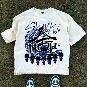 Może przedstawiać: Biała koszulka z czarno-niebieskim nadrukiem graficznym koreańskiego boysbandu Stray Kids. Grafika zawiera nazwę zespołu, stylizowany wz&oacute;r fali i grupę postaci z kresk&oacute;wek.