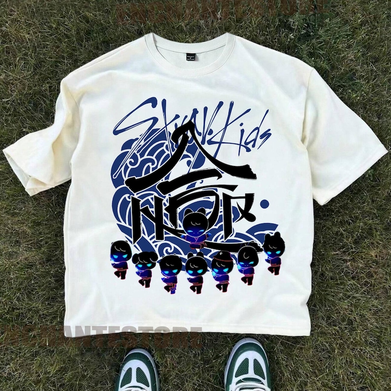 Może przedstawiać: Biała koszulka z czarno-niebieskim nadrukiem graficznym koreańskiego boysbandu Stray Kids. Grafika zawiera nazwę zespołu, stylizowany wz&oacute;r fali i grupę postaci z kresk&oacute;wek.