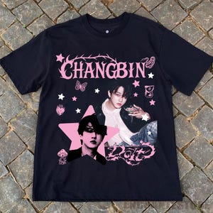 Pu&ograve; includere: T-shirt nera con grafica rosa raffigurante il nome "CHANGBIN" e la foto di una persona. Il design include stelle, una farfalla e la parola "Dote". La maglietta &egrave; realizzata in materiale morbido.