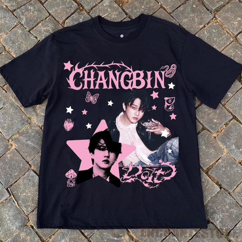 Pu&ograve; includere: T-shirt nera con grafica rosa raffigurante il nome "CHANGBIN" e la foto di una persona. Il design include stelle, una farfalla e la parola "Dote". La maglietta &egrave; realizzata in materiale morbido.