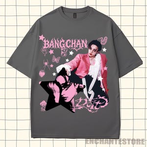 Pu&ograve; includere: T-shirt grigio con un design grafico rosa e nero. Il design include il testo "BANGCHAN", un ritratto, stelle e altri elementi decorativi. La maglietta &egrave; in stile casual.