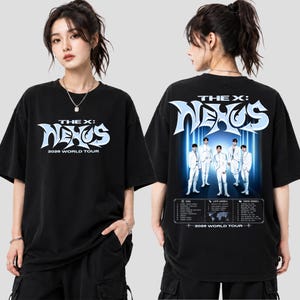May include: A black short-sleeve t-shirt featuring a graphic print. The front displays the text THE X: NEXUS 2026 WORLD TOUR in light blue and white. The back shows the same text above a photo of five people and a list of tour locations.