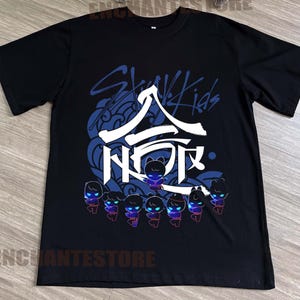 Może przedstawiać: Czarny T-shirt z niebiesko-białym nadrukiem graficznym z tekstem "Stray Kids" i stylizowanym japońskim znakiem. Grafika zawiera r&oacute;wnież osiem małych postaci z kresk&oacute;wek w kolorze czarnym i niebieskim.