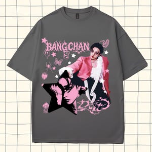 Pode incluir: Uma camiseta cinza com um design gr&aacute;fico rosa com o texto "BANGCHAN" e um retrato de uma pessoa com um casaco rosa. O design inclui estrelas, borboletas e outros elementos decorativos.
