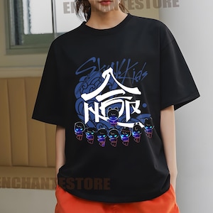 Może przedstawiać: Czarny T-shirt z nadrukiem graficznym koreańskiego boysbandu Stray Kids. Grafika przedstawia nazwę zespołu w języku koreańskim i angielskim, a także stylizowany wizerunek członk&oacute;w zespołu.