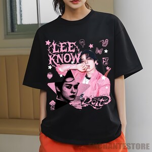 Pu&ograve; includere: T-shirt nero con un design grafico rosa con la scritta "LEE KNOW" e due ritratti di una persona. Il design include stelle, una farfalla e altri elementi decorativi. La persona nei ritratti indossa una giacca rosa.