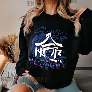 Może przedstawiać: Czarna bluza z nadrukiem graficznym koreańskiego boysbandu Stray Kids. Grafika zawiera nazwę zespołu w języku koreańskim i angielskim, a także stylizowany wz&oacute;r fali i postacie z kresk&oacute;wek.