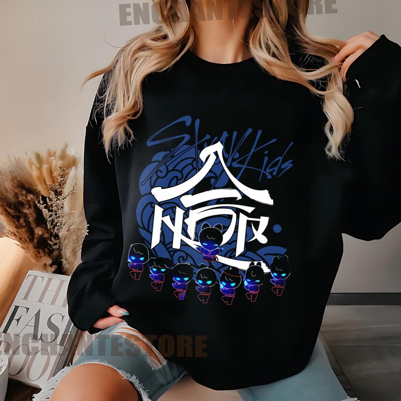 Może przedstawiać: Czarna bluza z nadrukiem graficznym koreańskiego boysbandu Stray Kids. Grafika zawiera nazwę zespołu w języku koreańskim i angielskim, a także stylizowany wz&oacute;r fali i postacie z kresk&oacute;wek.