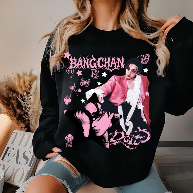 Pode incluir: Camisola preta com um gr&aacute;fico rosa de uma pessoa com um blazer rosa, com o texto "BANGCHAN" e "ZONE". O design inclui estrelas, uma aranha e uma borboleta. A camisola &eacute; de estilo casual.