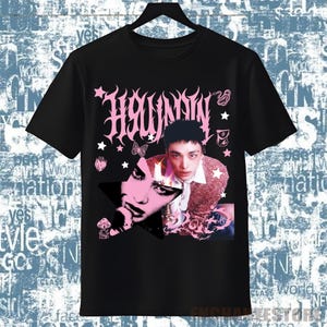 Pu&ograve; includere: T-shirt nera con un design grafico rosa. Il design presenta la parola "Hwaunnn" in un font stilizzato, un ritratto di una persona, stelle e altri elementi decorativi. La maglietta &egrave; appesa a una gruccia nera.