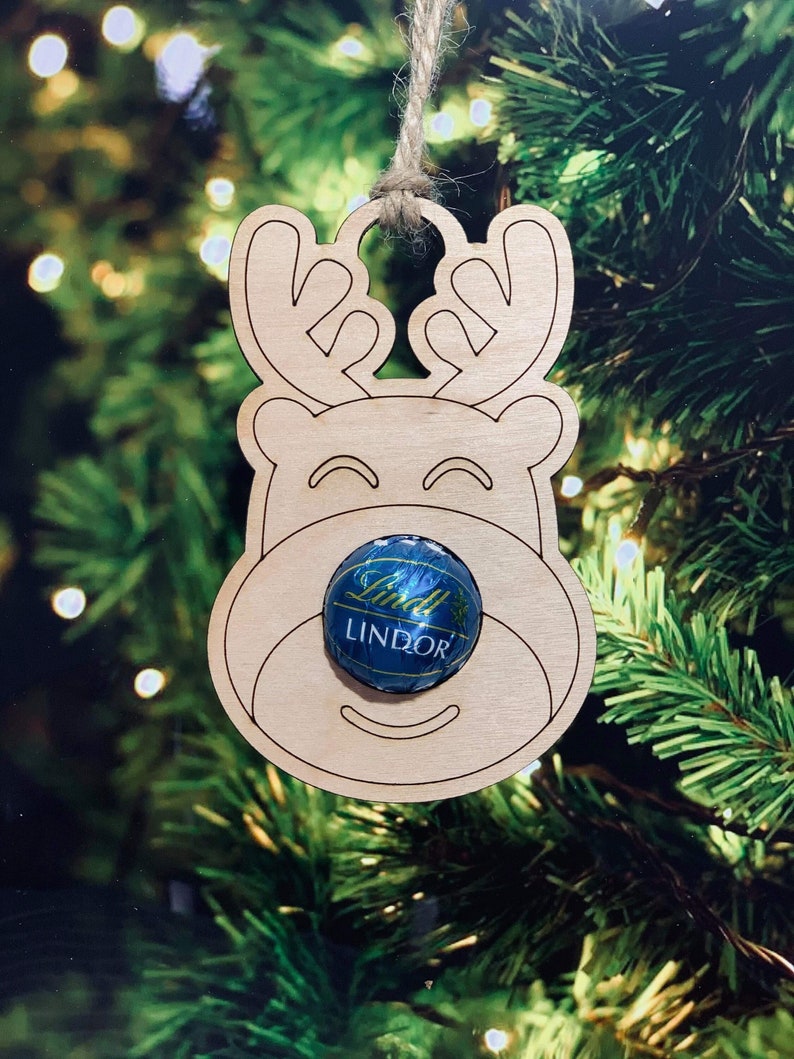 K&ouml;nnte beinhalten: Weihnachtsbaumschmuck aus Holz in Form eines Rentiers mit einem Lindor-Schokoladentaler in der Nase. Der Schmuck h&auml;ngt an einem Weihnachtsbaum.