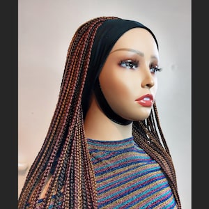 以下が含まれることがあります： 長い茶色と金のボックスブレードをしたマネキンヘッド。ブレードは黒いヘッドバンドでスタイリングされています。マネキンはストライプのシャツを着ています。