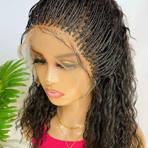 May include: A black braided wig with wavy, curly hair. The wig features a lace front and is styled on a mannequin head. The mannequin is wearing a pink top and a gold necklace with a pendant. The wig is a dark shade of black.