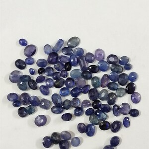 May include: A collection of loose blue sapphire gemstones in various sizes and shapes. The gemstones are oval, round, and rectangular. They are all a deep blue colour with a slight purple hue.