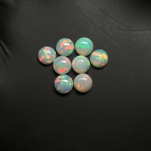 May include: Eight round, white opal cabochons with iridescent rainbow flashes. The opals are arranged in a circular pattern on a black surface.