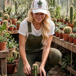 Könnte beinhalten: Eine weiße Baseballkappe mit einer grünen Kaktusgrafik in einem rosa Topf und einer perlenartigen Verzierung entlang des Schirms. Die Kappe verfügt über eine strukturierte Krone und einen gebogenen Schirm in einem schlichten Design.