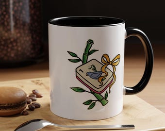 May include: A white ceramic mug with a black interior and handle. The exterior features a graphic of a mahjong tile with a blue bird illustration, a number one, green bamboo stalks, and a gold ribbon tied around the tile.