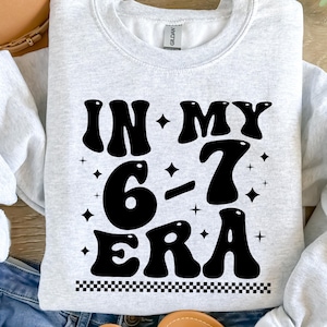 以下が含まれることがあります： レトロなフォントで「IN MY 6-7 ERA」というテキストがプリントされたライトグレーのスウェットシャツ。黒いテキストには星のアクセントが付いています。テキストの下部には、白と黒の市松模様があります。