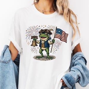 Könnte beinhalten: Weißes T-Shirt mit einer Cartoon-Froschfigur in Kolonialkleidung, die eine US-Flagge und eine Glocke hält, mit Feuerwerk im Hintergrund. Das Design verwendet grüne, blaue, braune und rote Farben.