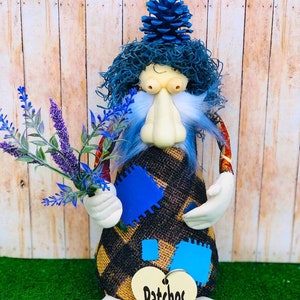 Puede incluir: Una muñeca artesanal caprichosa con un sombrero de piña azul, cabello azul y un atuendo de retazos. La muñeca sostiene un ramo de lavanda y tiene una etiqueta en forma de corazón que dice "Patches".