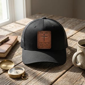Può includere: Cappellino da camionista nero con retro in rete e toppa in pelle. La toppa presenta una croce e le parole "THE WAY, THE TRUTH, THE LIFE". Il cappellino è su una superficie di legno.
