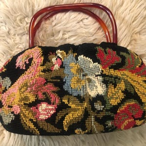 Puede incluir: Un bolso negro vintage con un diseño floral de punto de aguja en rosa, amarillo, azul y rojo. El bolso tiene dos asas curvas de plástico marrón.