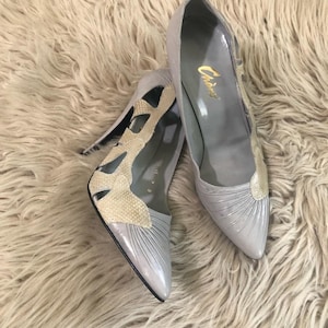 Peut inclure: Une paire d'escarpins gris clair à bout pointu avec une superposition beige texturée ajourée sur les côtés et un design plissé sur le bout. La doublure intérieure est grise avec le nom de marque Chino imprimé en lettres dorées sur la semelle.