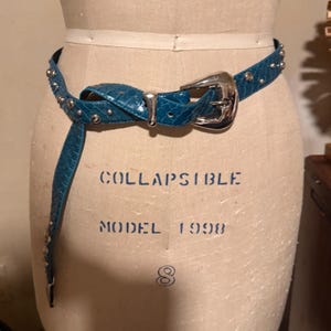May include: A teal belt with a textured snakeskin pattern, featuring silver-toned metal studs along the strap and a polished silver-toned metal buckle. The belt is wrapped around a beige dress form that displays the text COLLAPSIBLE MODEL 1998.
