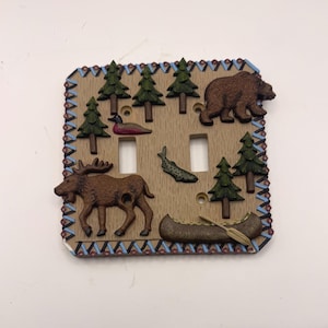 May include: A square light switch wall plate featuring a tan wood-grain finish with raised relief figures of a brown bear, a moose, a canoe with a paddle, a fish, and several green pine trees, bordered by a blue and red geometric pattern.