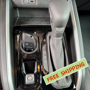 May include: Close-up of a black car's center console with a leather-wrapped gear shifter and a black electronic parking brake button. The gear shifter has the letters "P", "R", "N", "D", and "L" printed on it. The console also has a button with a "P" symbol for the parking brake.