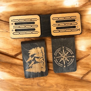 May include: A wooden cribbage board set with two playing boards and two card holders. The playing boards are light brown with black accents and drilled holes. The card holders are black with etched designs, one with a compass and the other with a mountain scene.
