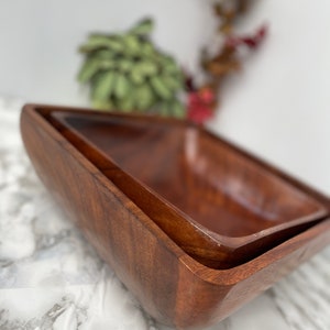 Puede incluir: Dos cuencos de madera, uno anidado dentro del otro, están hechos de madera marrón oscura. Los cuencos son de forma cuadrada y tienen un acabado liso y pulido.