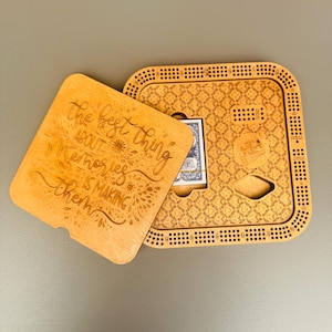 May include: A wooden cribbage board with a decorative lid. The lid is engraved with the phrase "The best thing about memories is making them." The board has a patterned design and holes for pegs. A deck of cards is visible.