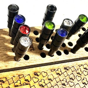 Peut inclure: Un plateau de jeu en bois avec une grille de trous et une rangée de chevilles surmontées de gemmes colorées. Les gemmes sont noires, bleues, rouges, vertes et transparentes. Le plateau comporte une section avec un motif de cœurs, de diamants, de trèfles et de piques.