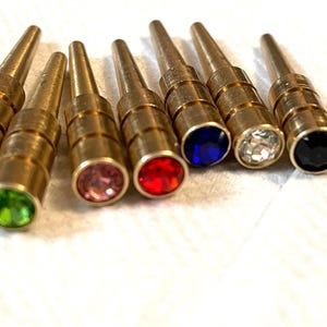 Peut inclure: Un ensemble de huit épingles métalliques cylindriques couleur laiton, chacune surmontée d'une pierre précieuse à facettes. Les pierres sont de différentes couleurs, dont transparent, vert, rose, rouge, bleu et noir, alignées.