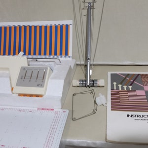 May include: A white and orange striped fabric sample, a white plastic loom with a control panel labeled "YC-3", a metal loom with a white spool, a white plastic frame, and an instruction book for an "Automatic Yarn Changer YC-3" loom.
