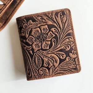 Może przedstawiać: Brązowy skórzany portfel z wytłoczonym kwiatowym wzorem. Portfel jest prostokątny i ma szczegółowy wzór kwiatów i liści. Na zdjęciu widoczny jest również otwarty portfel.