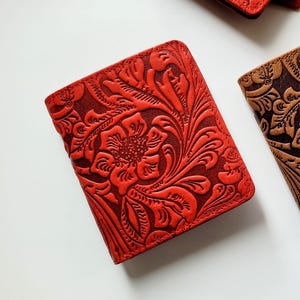 May include: Close-up of several leather notebooks with floral embossed designs. One notebook is red, another is brown, and a third is red. The notebooks are rectangular and have a detailed, textured appearance.
