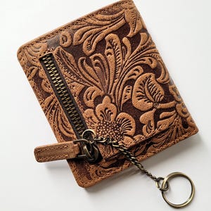 May include: Brown leather wallet with a floral embossed design. The wallet has a zipper closure and a key ring attached with a chain. The wallet is rectangular and appears to be new.