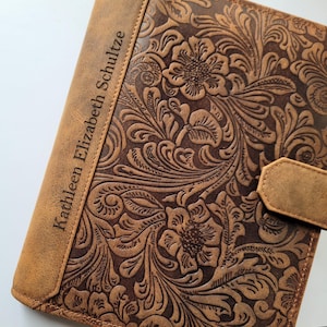 Könnte beinhalten: Ein braunes Lederjournal mit geprägtem Blumenmuster. Der Name "Kathleen Elizabeth Schultze" ist vertikal auf dem Buchrücken eingraviert. Ein Lederriemen mit Druckknopfverschluss ist sichtbar.