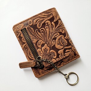 May include: Brown leather wallet with a floral embossed design. The wallet has a zipper closure and a key ring attached with a chain. The wallet is rectangular and appears to be new.